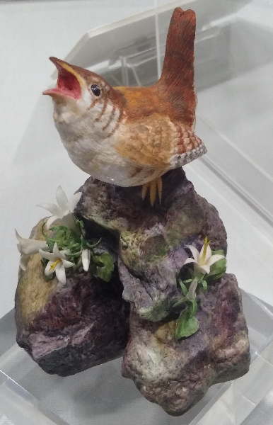 Decorative ornament of a bird with its beak open, standing on a rock with small white flowers